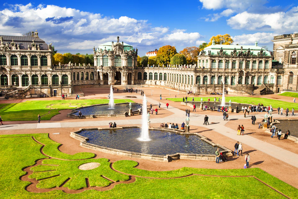Ornate Baroque palace with fountains and gardens, people walking and sitting, under a blue sky with scattered clouds.