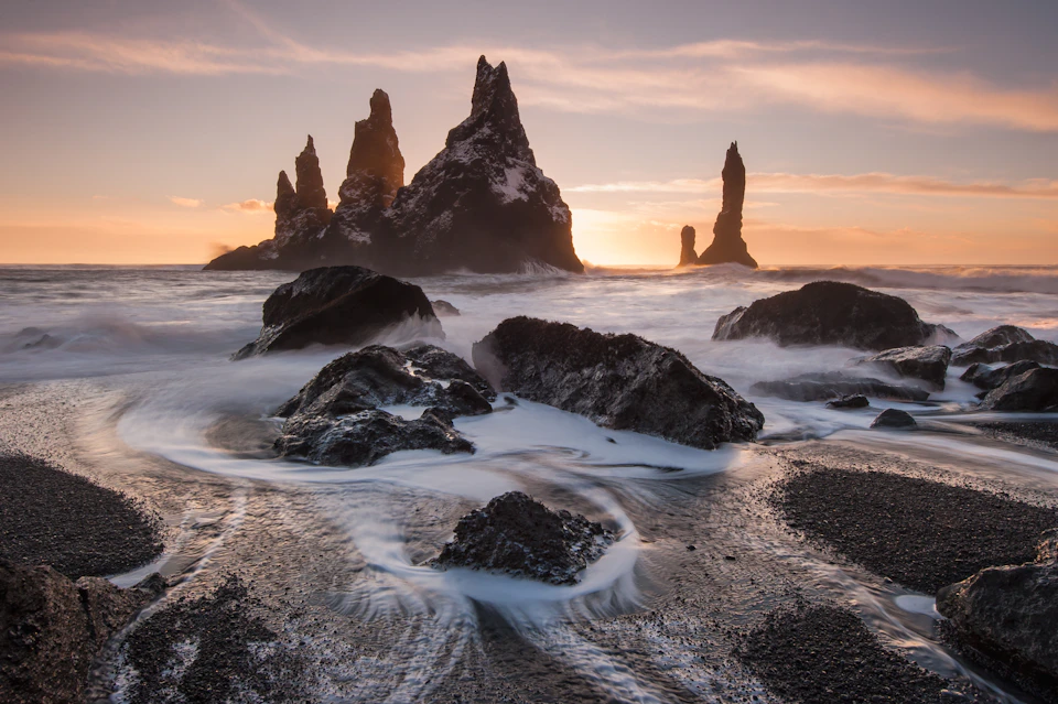 Dramatic sea stacks at sunset stand in rough waters, with waves swirling around dark rocks on a black sand beach.