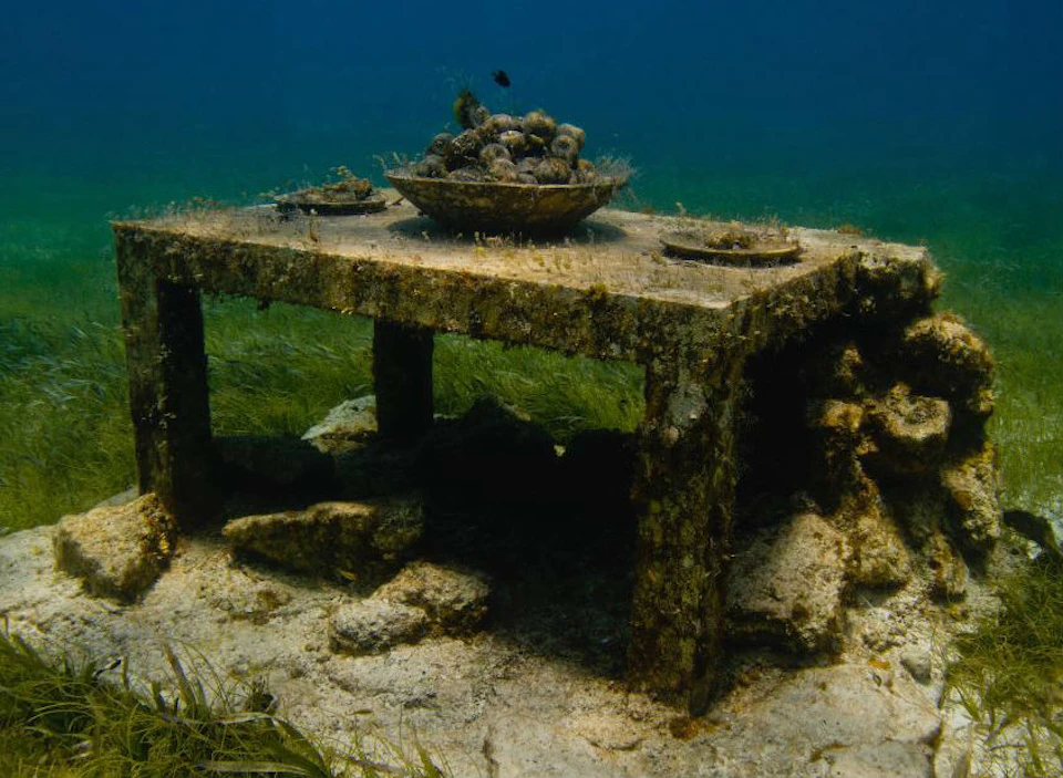 An underwater table covered in algae with a large bowl of seashells and two smaller plates on its surface.