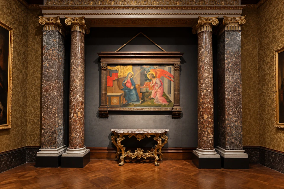 A framed artwork depicting a religious scene hangs on a dark wall between two marble columns with a decorative table below.