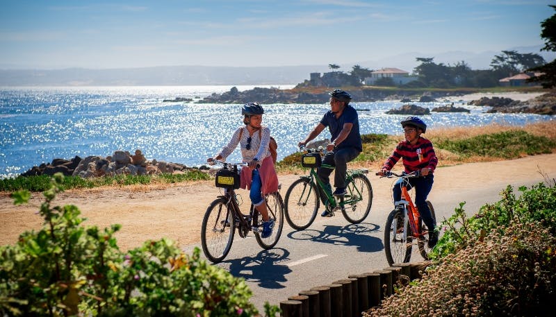 Three people ride bicycles on a coastal path with the ocean and houses in the background.