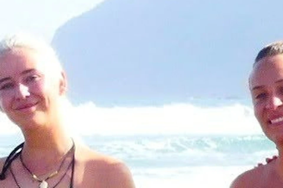 Six people in swimsuits and wetsuit pants stand on a beach with mountains in the background.