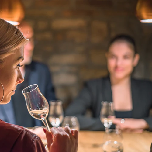 En kvinna med kort blont hår undersöker ett glas vin, sittande med två suddiga individer i bakgrunden.