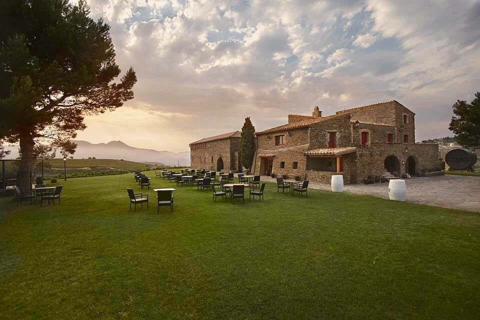 Stone buildings and several sets of tables and chairs on a grassy area with mountains in the background under a cloudy sky.
