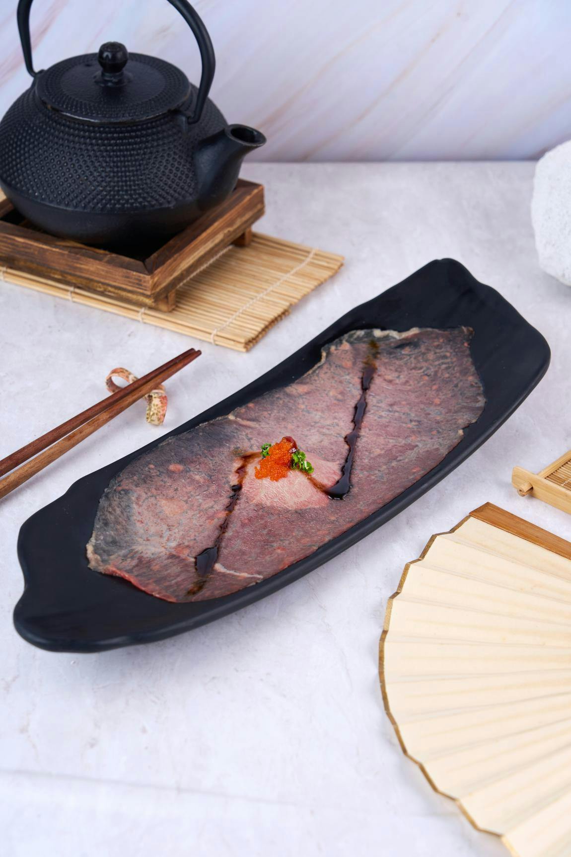 Thinly sliced raw meat garnished with roe and herbs on a black plate, surrounded by chopsticks and a fan on a table.