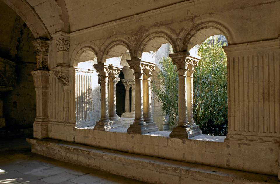 A stone corridor with ornate arches and columns overlooks a sunlit courtyard with a large leafy plant.