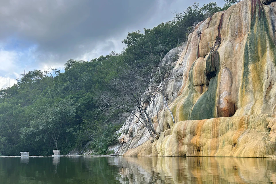 A natural rock formation with mineral deposits next to a calm body of water, surrounded by green foliage under a cloudy sky.