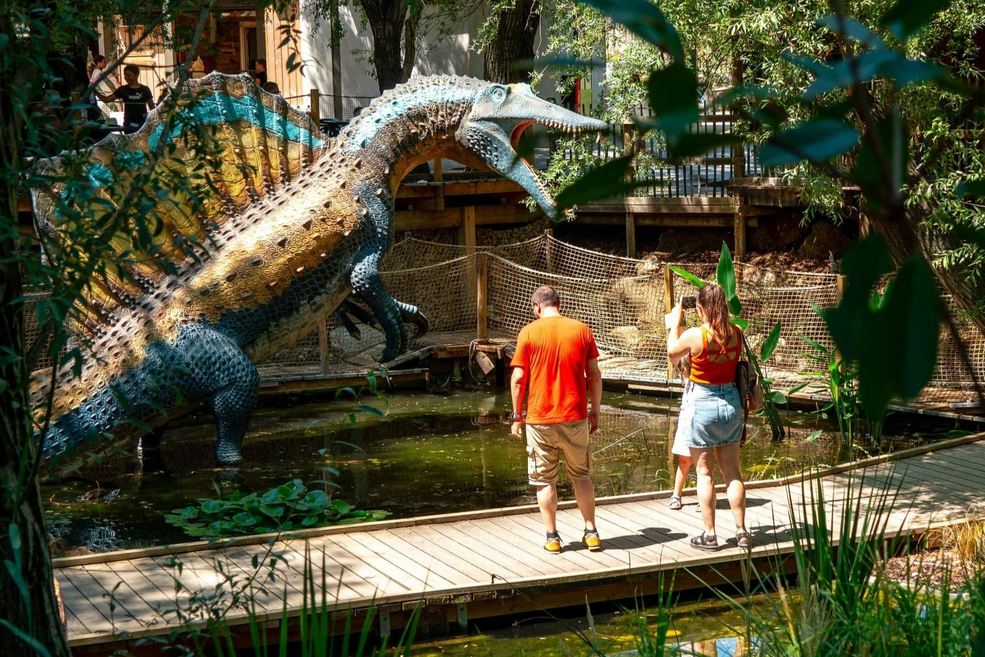 Una gran escultura de dinosaure prop d'un estany, amb dues persones en una passarel·la de fusta observant-la i fent fotos.