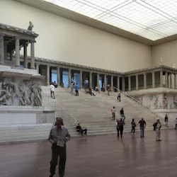 Besökare i ett museum med ett stort altare i antik grekisk stil och invecklade skulpturer, stående och sittande runt utställningen.