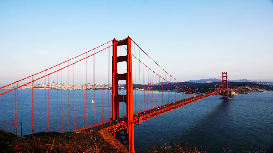 The Golden Gate Bridge spanning across the water with a sailboat in the distance and a clear blue sky.