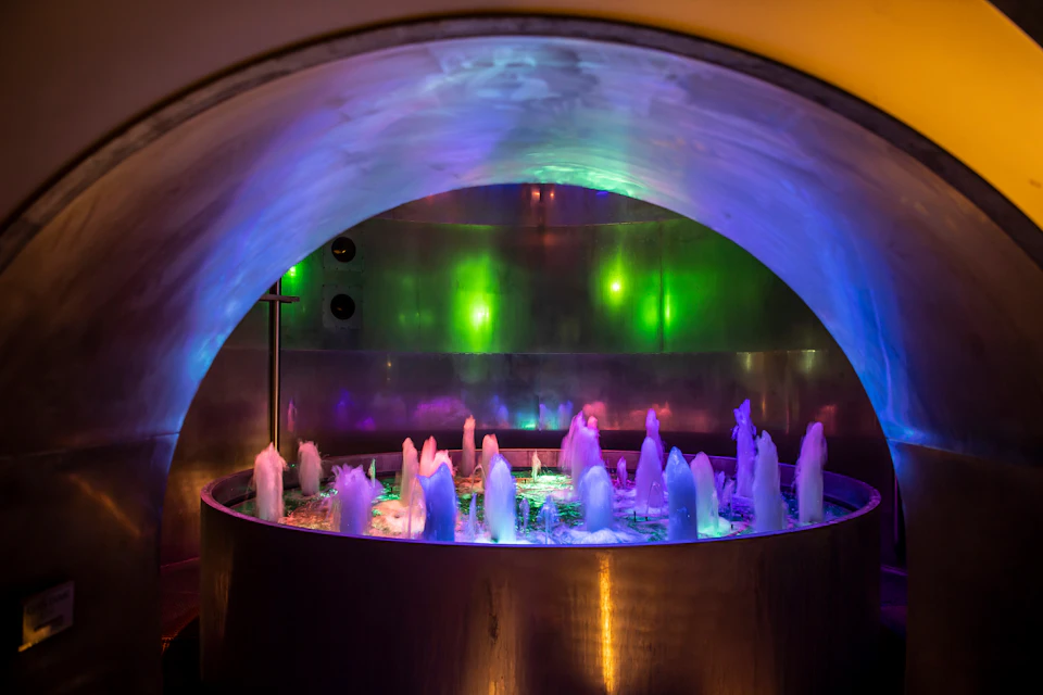 A circular indoor fountain with multicolored lights illuminating water jets against a metallic backdrop.