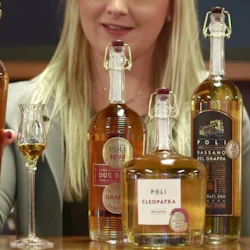 En kvinna med långt blont hår visar fem flaskor Poli grappa och ett fyllt glas på en träyta.