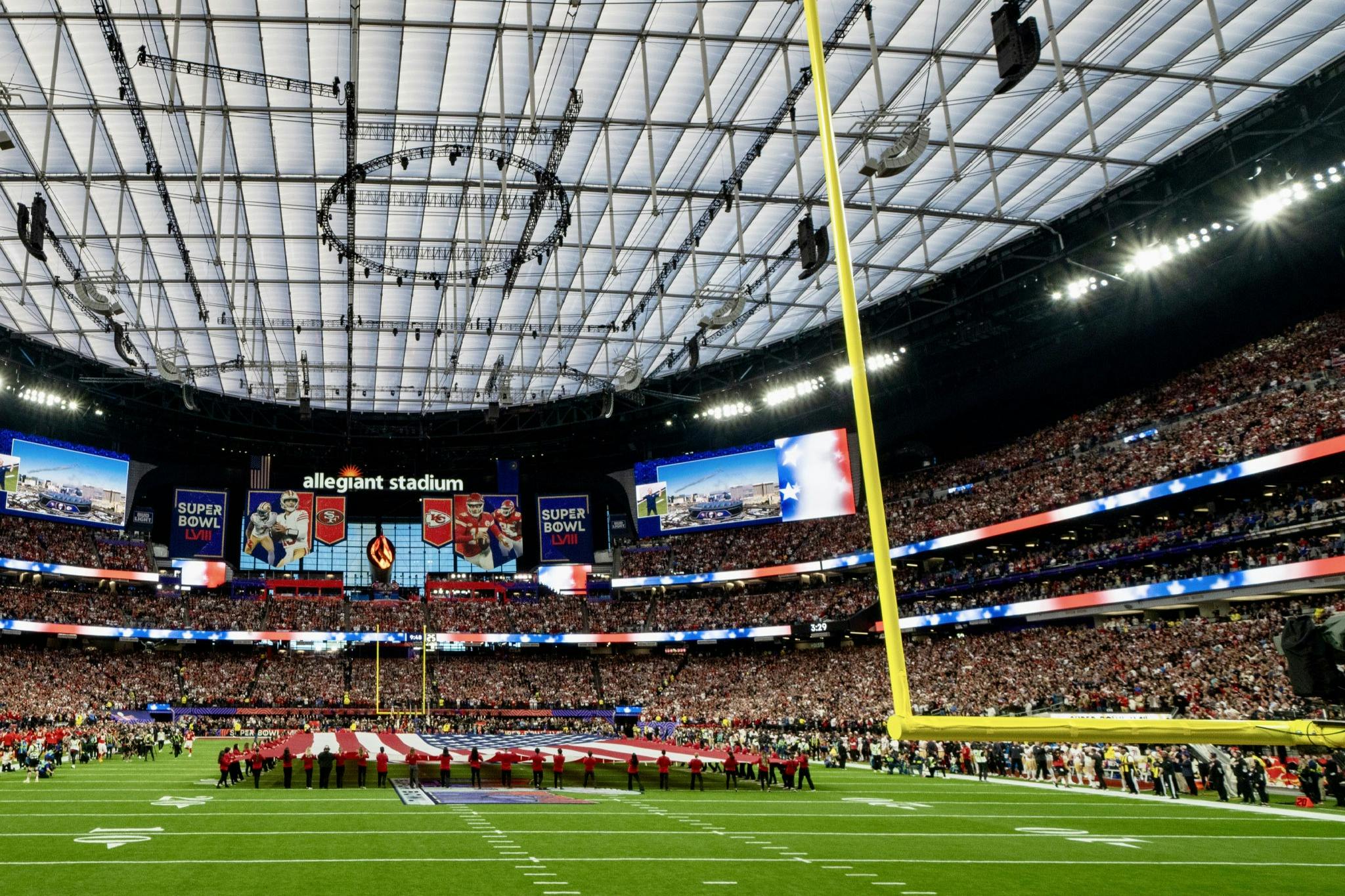A football stadium with a large American flag displayed on the field during a pregame ceremony, surrounded by a crowd.