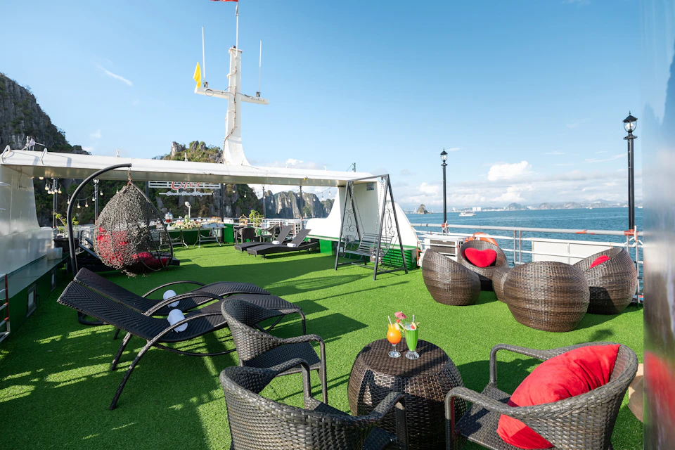 Outdoor lounge area on a ship with wicker furniture, drinks on tables, hanging chairs, and a clear blue sky with coastal scenery.