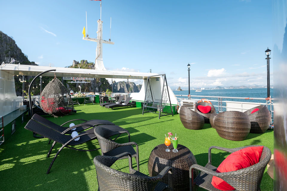 Outdoor lounge area on a ship with wicker furniture, drinks on tables, hanging chairs, and a clear blue sky with coastal scenery.