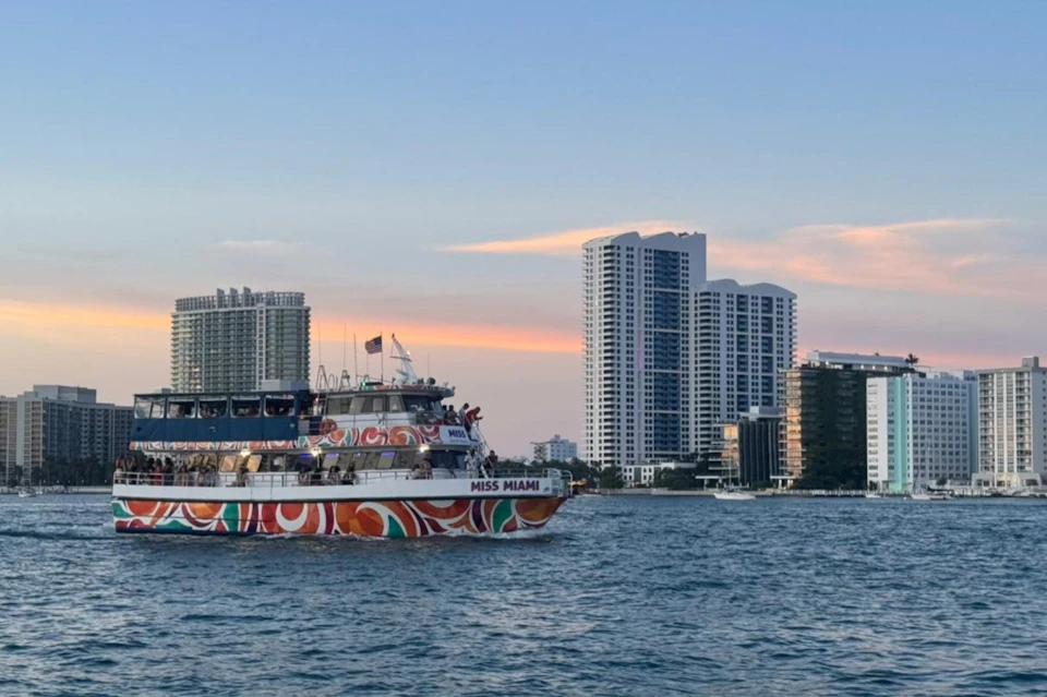 A colorful boat named 'Miss Miami' on water, with tall buildings in the background under a soft evening sky.