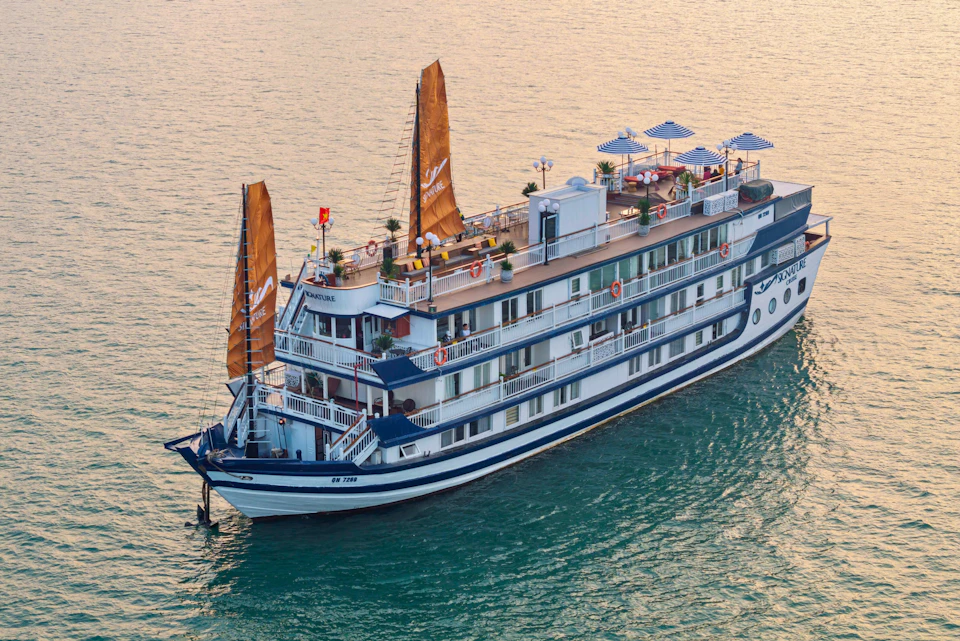 A multi-deck white cruise ship with orange sails and outdoor seating sails on calm water.