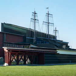 En stor modern byggnad med skeppsmaster som sträcker sig från taket och en svensk flagga på toppen, ställd mot en gräsbevuxen förgrund.