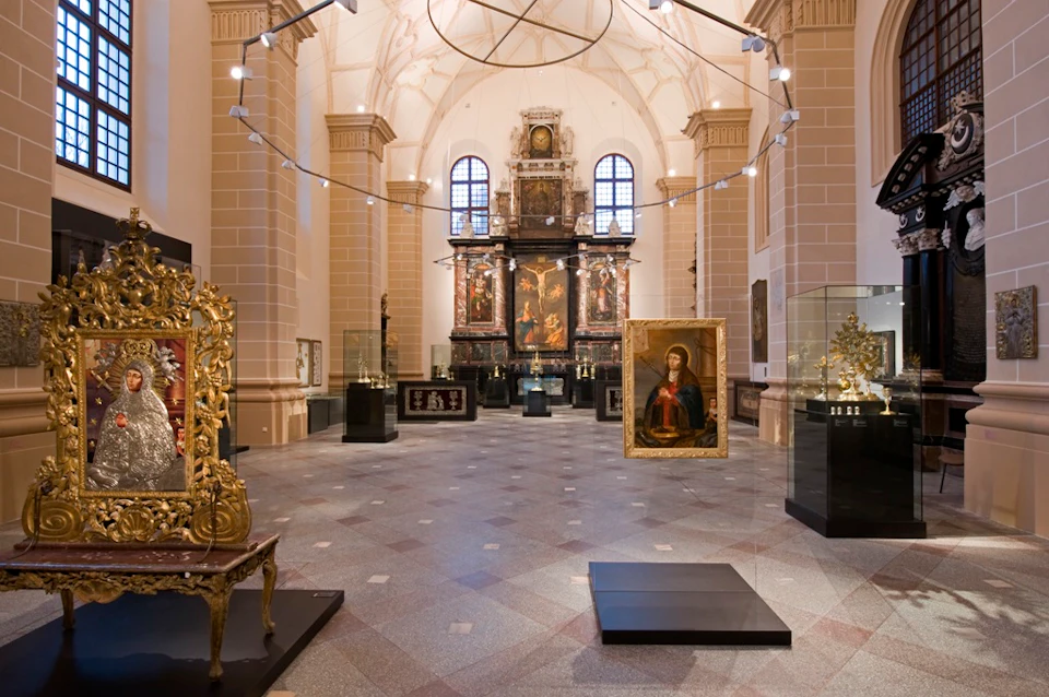 A spacious art gallery with various framed paintings, sculptures, and display cases under a high vaulted ceiling with arched windows.