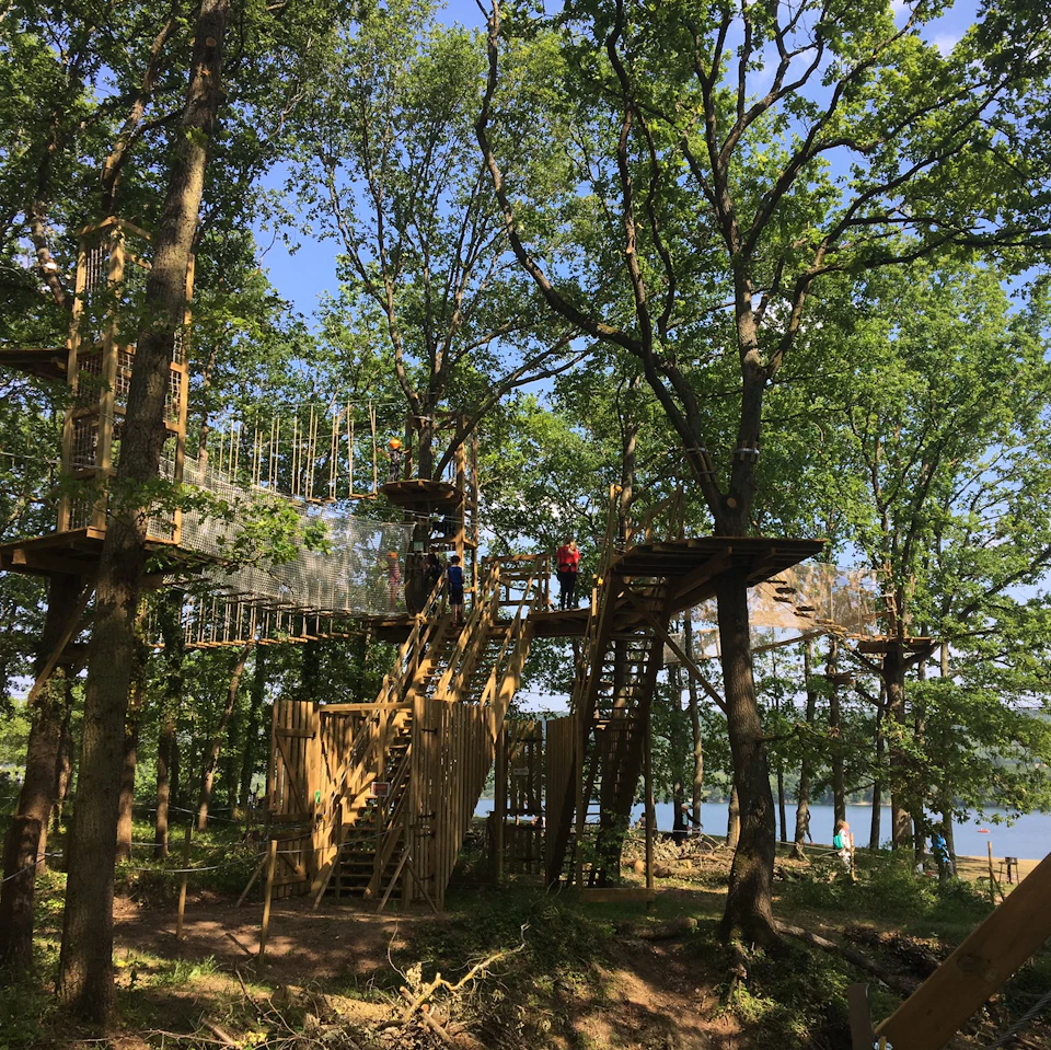 Treehouse adventure park with wooden platforms, bridges, and ladders among tall trees; people are walking on the structures.