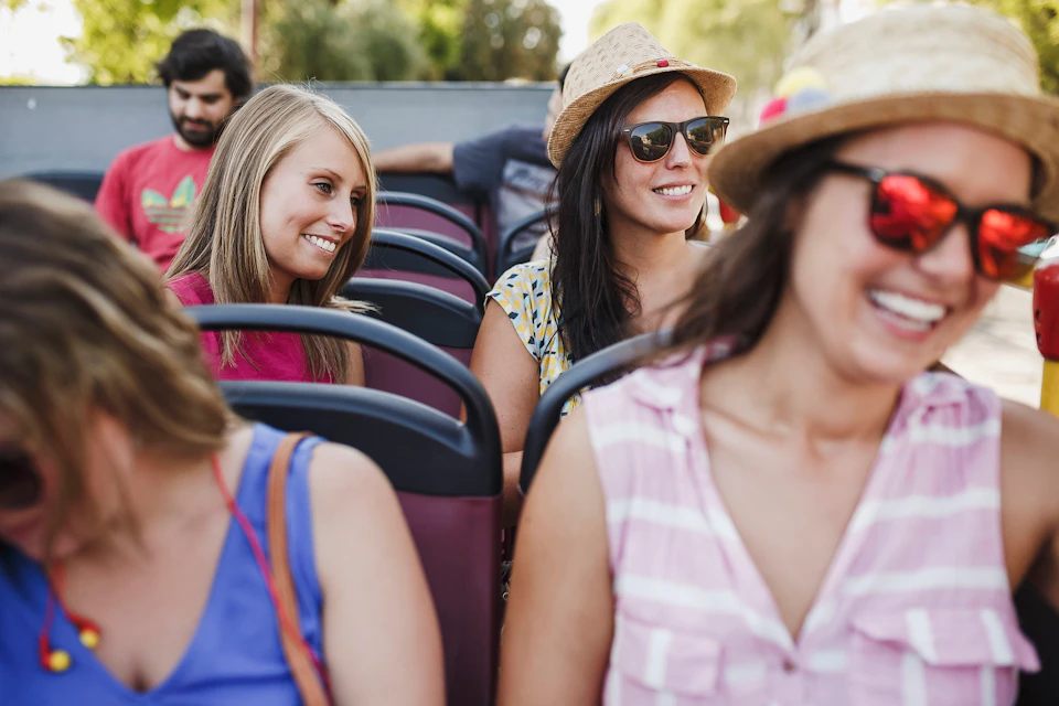 Four women wearing sunglasses and hats smiling while seated on a bus or open-air vehicle.
