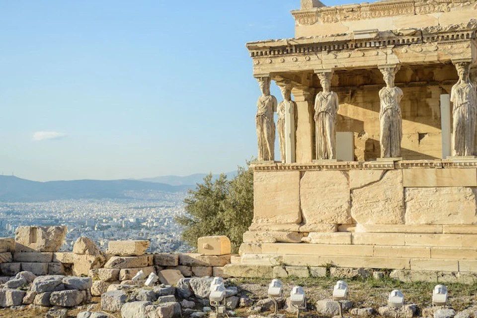Ancient Greek temple with caryatid columns, overlooking a distant cityscape under a clear blue sky.
