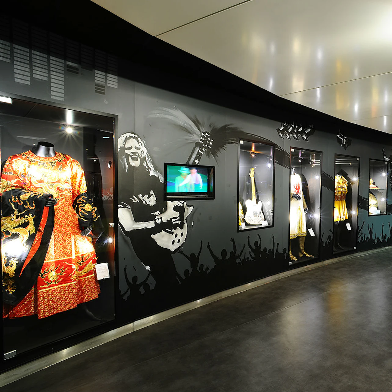A museum exhibit wall showcasing rock memorabilia, with costumes, guitars, and a video display.