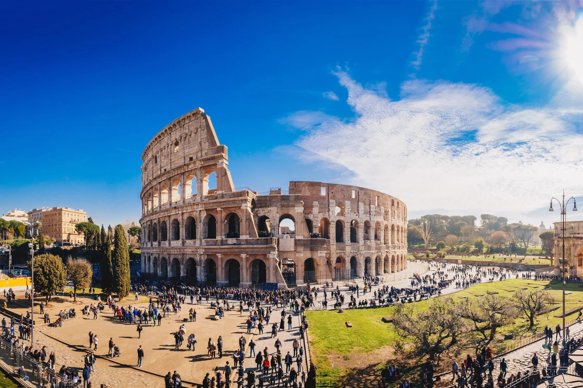The Colosseum in Rome under a clear blue sky, with many tourists walking around the surrounding area.