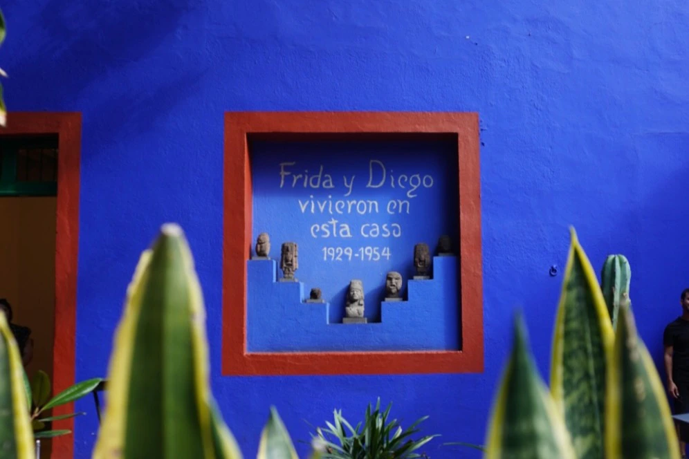 Blue wall inset with clay figurines and text: "Frida y Diego vivieron en esta casa 1929-1954," framed by plants.