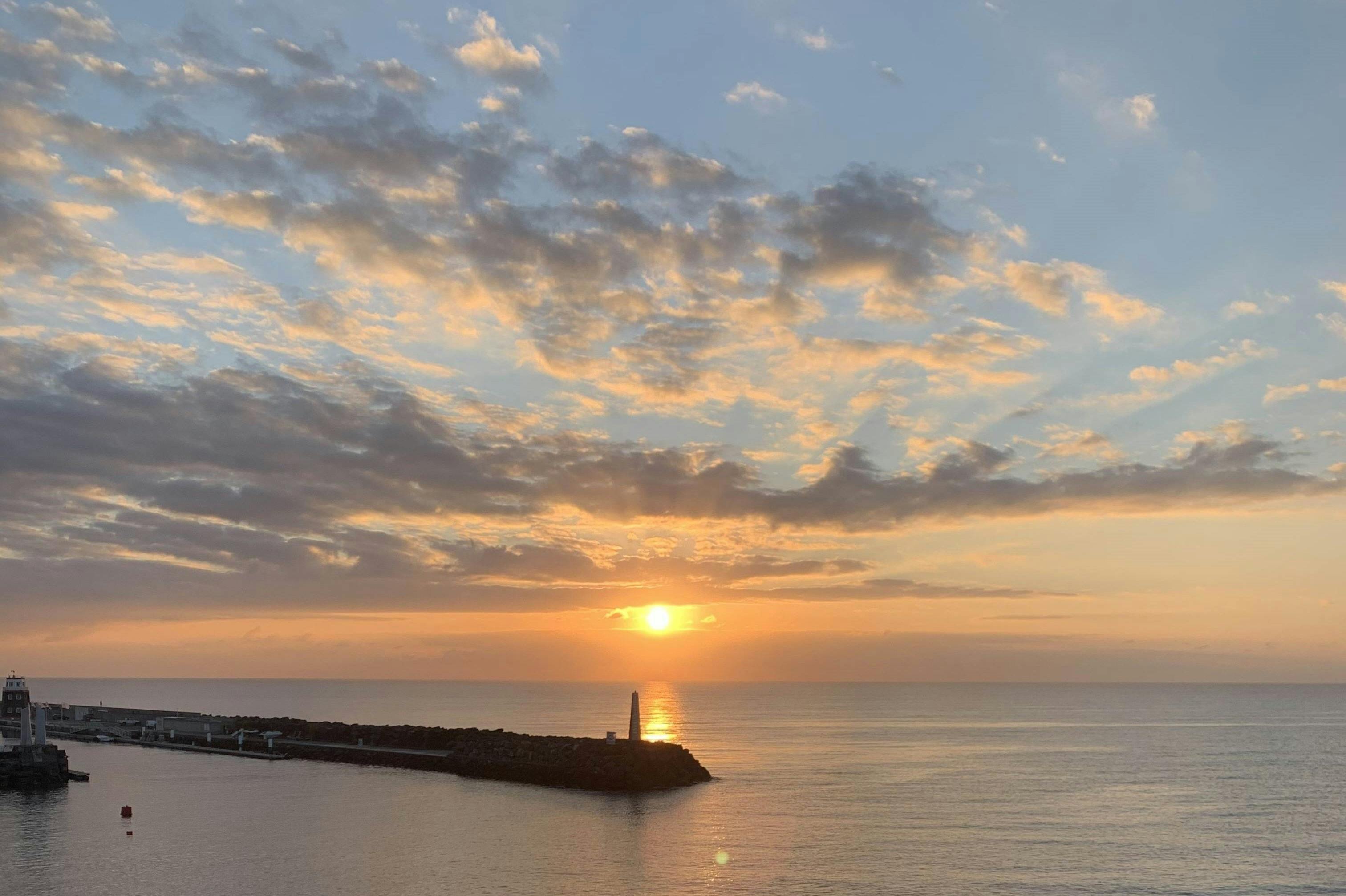 석양을 반사하는 흩날리는 구름으로 가득한 하늘 아래, 바위 부두의 등대와 함께 잔잔한 바다 위로 석양을 감상해보세요.