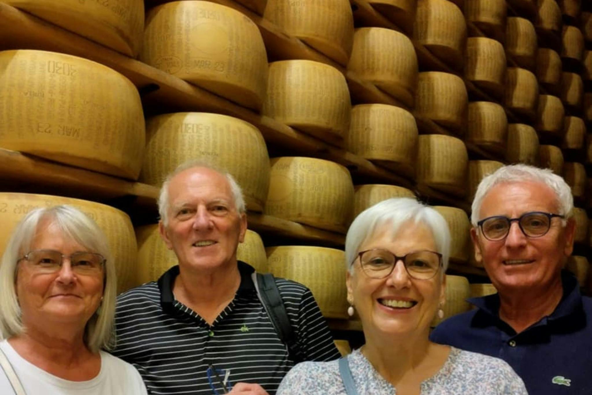 Tre anziani sorridono davanti a pile di forme di formaggio.