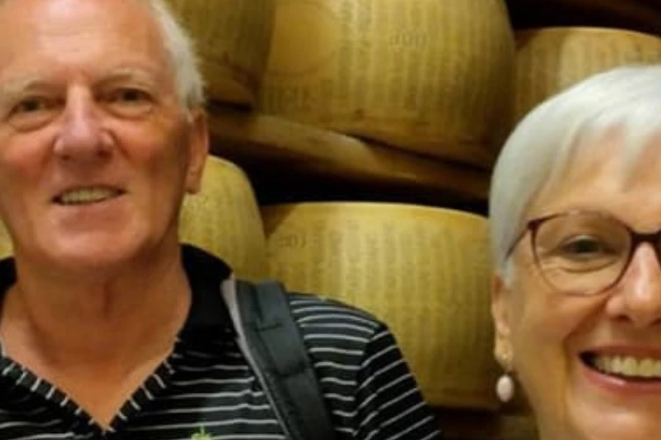 Three elderly individuals are smiling in front of stacks of cheese wheels.