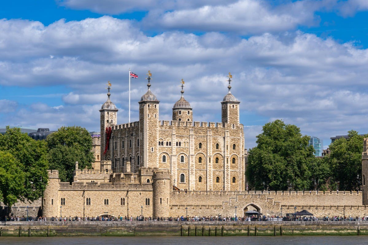 A historic stone castle with towers and a flag, surrounded by trees, under a partly cloudy blue sky.