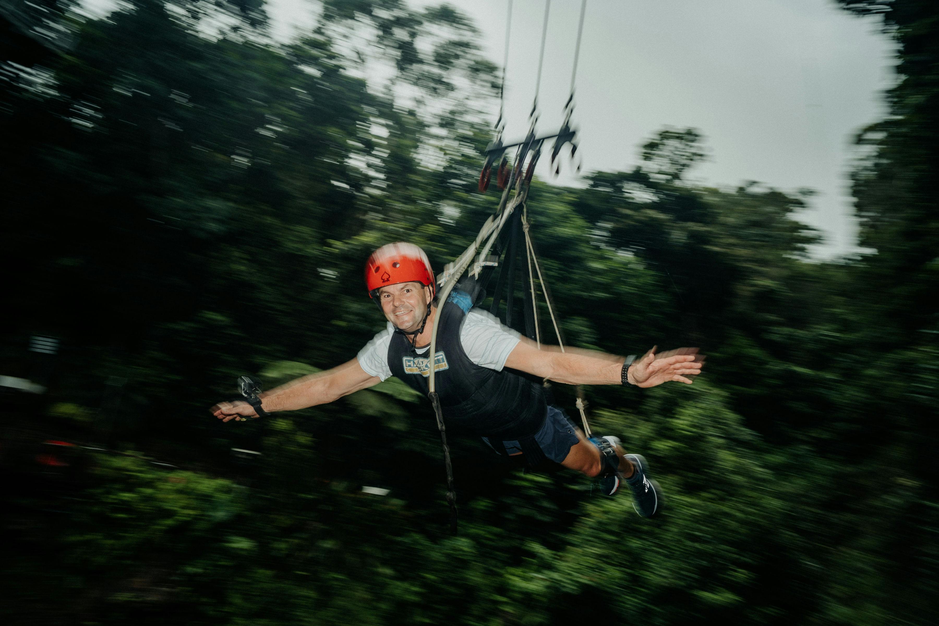 一个戴着红色头盔和马具的人悬挂在郁郁葱葱的绿色森林上空。 它们看起来像是高空滑索。