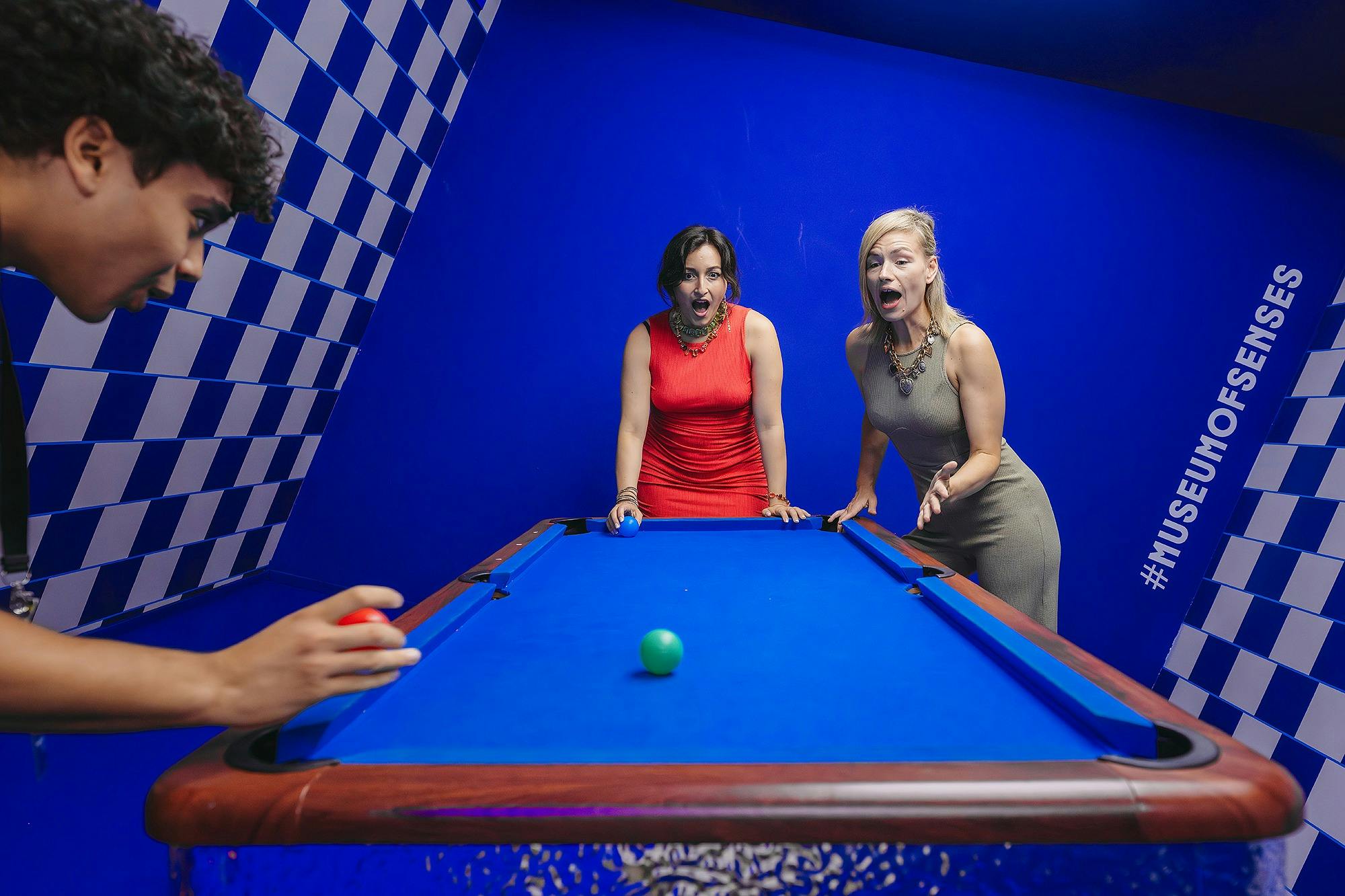 Tre personer interagerar med ett blått biljardbord i ett rum med blå och vita rutiga väggar, och verkar förvånade och roade.