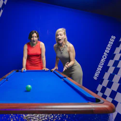 Tre personer interagerar med ett blått biljardbord i ett rum med blå och vita rutiga väggar, och verkar förvånade och roade.