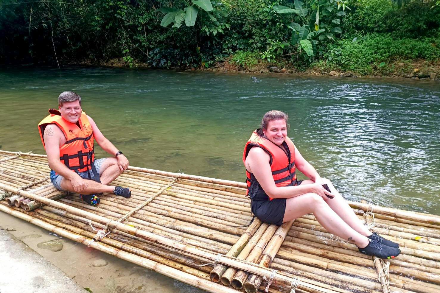 A person wearing a life jacket sits on a bamboo raft on a river, surrounded by lush greenery.