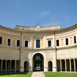 Böjd historisk byggnad med välvda ingångar, pelare och en klocka på toppen, placerad under en klarblå himmel.