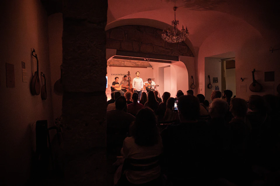 A small live performance in a dimly lit room with a singer and musicians on stage, and an audience seated in front.