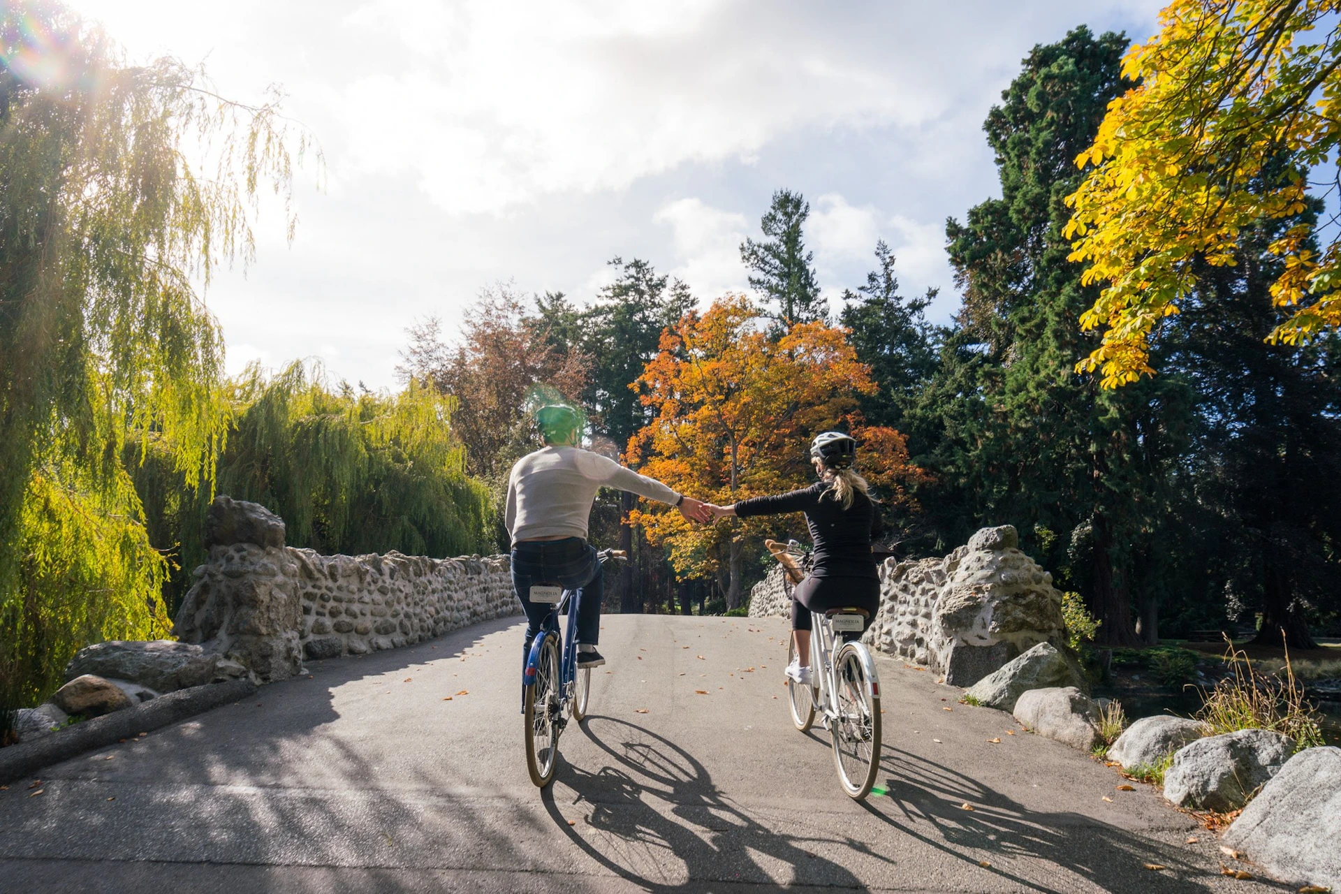Two people cycling on a paved trail in a park with autumn-colored trees, holding hands, surrounded by stone walls.