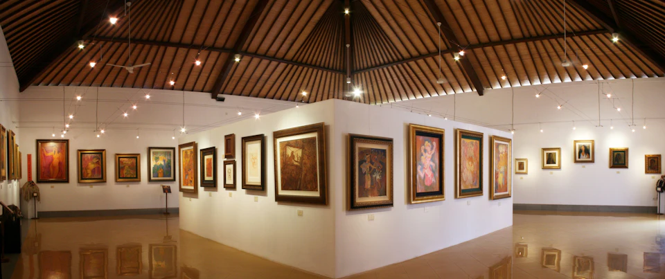Art gallery with framed paintings on white walls, under a wooden vaulted ceiling with spotlights.