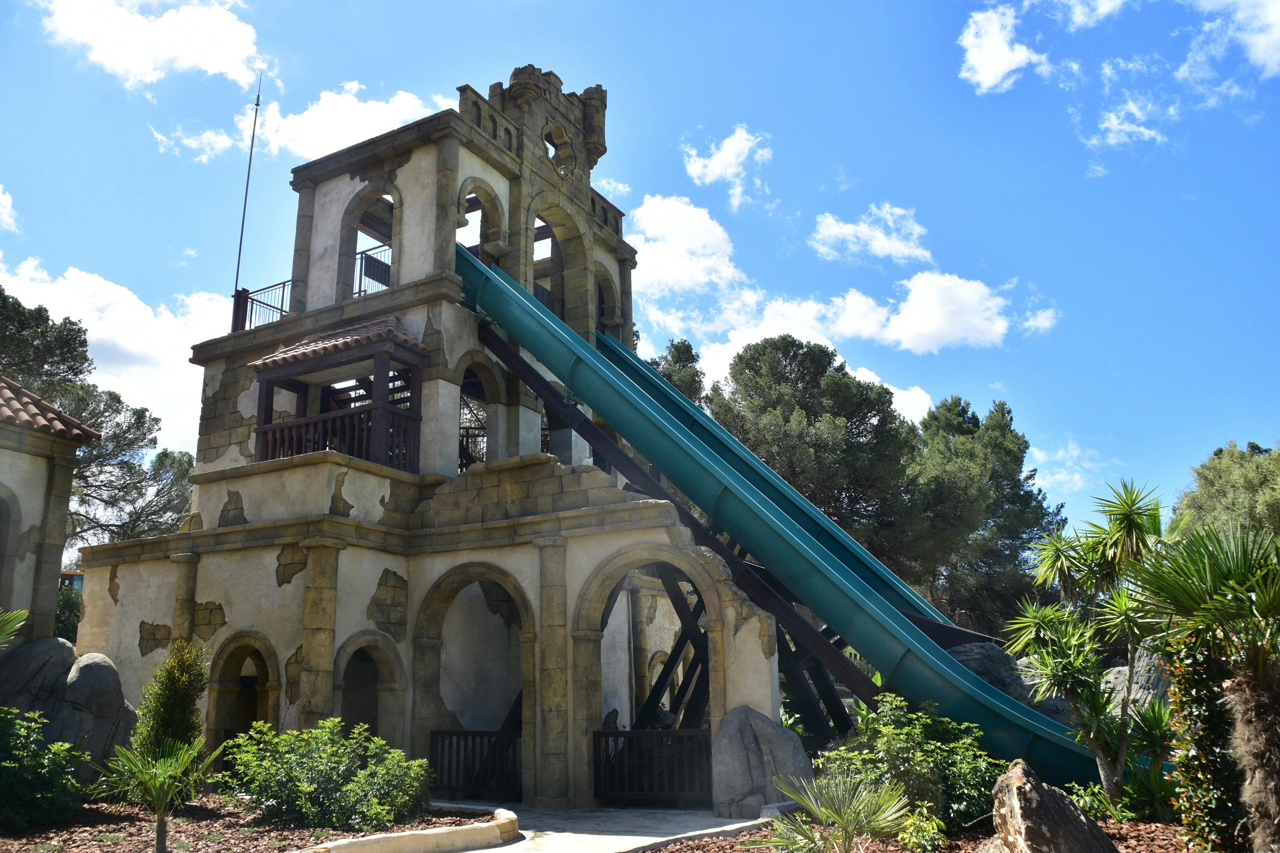A tall stone structure resembling a bell tower with a long blue slide attached, surrounded by trees and greenery under a blue sky.