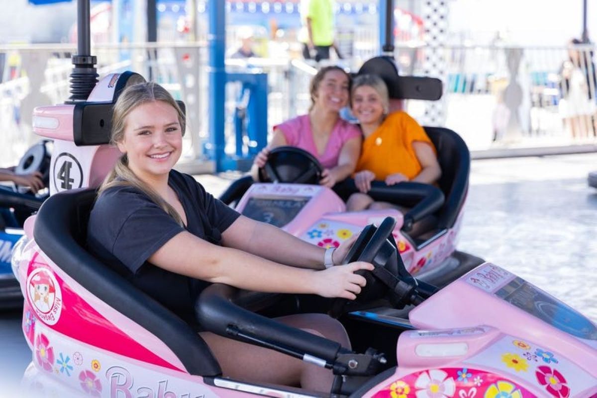 Three women smiling in pink and white go-karts at an amusement park. The woman in the foreground is driving.