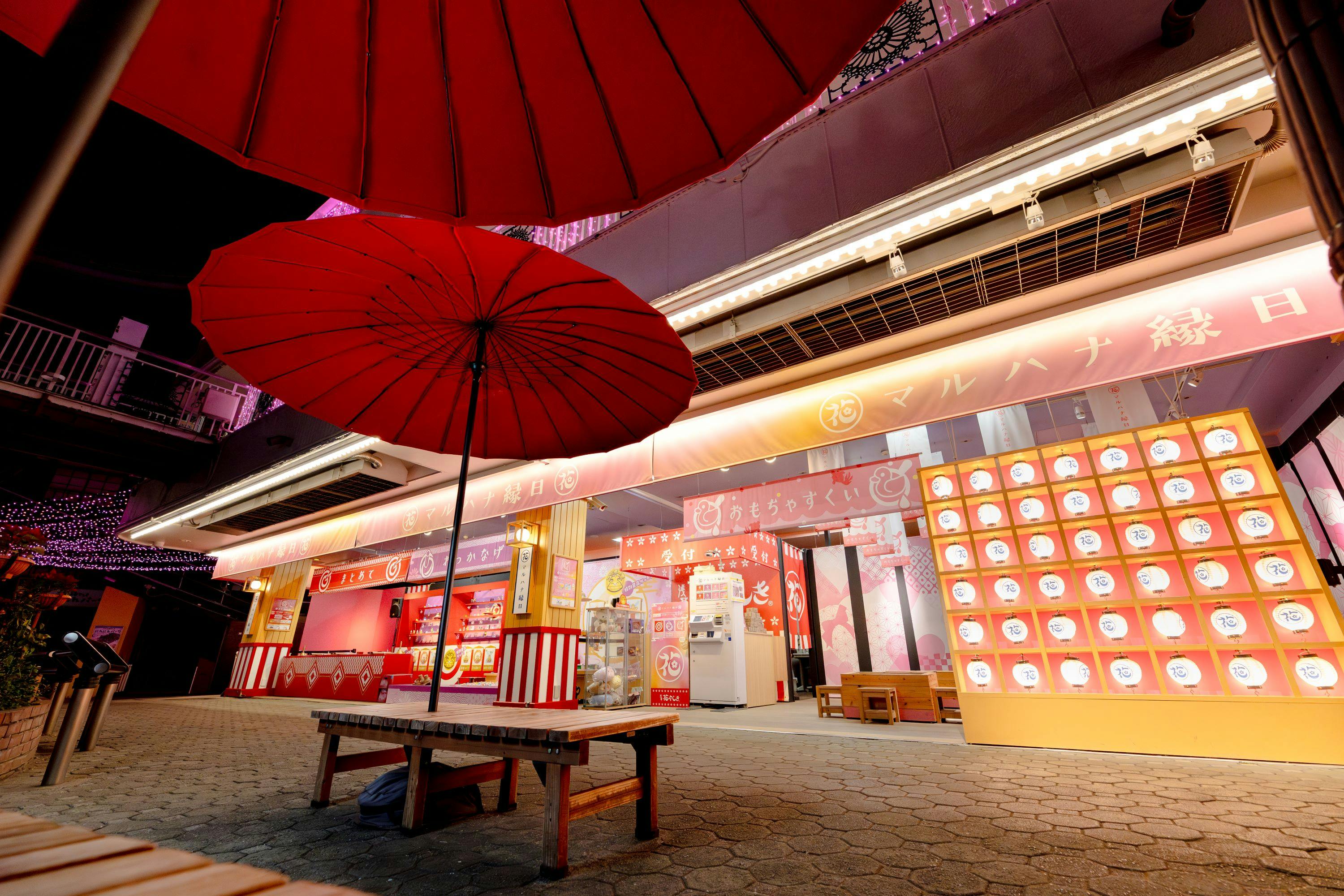 An outdoor seating area with red umbrellas in front of a brightly lit storefront with colorful signage.