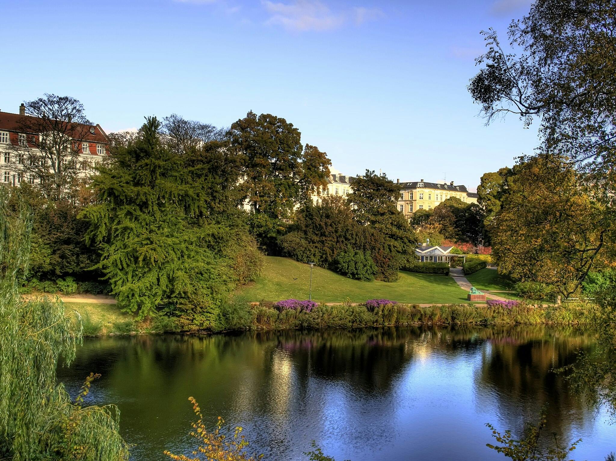 Scéna v parku s klidným rybníkem, svěží zelení, barevnými květinami a budovami v pozadí pod jasně modrou oblohou.