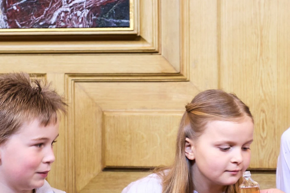 A group of children standing indoors facing a woman who is showing them an object, in a room with wooden paneling.