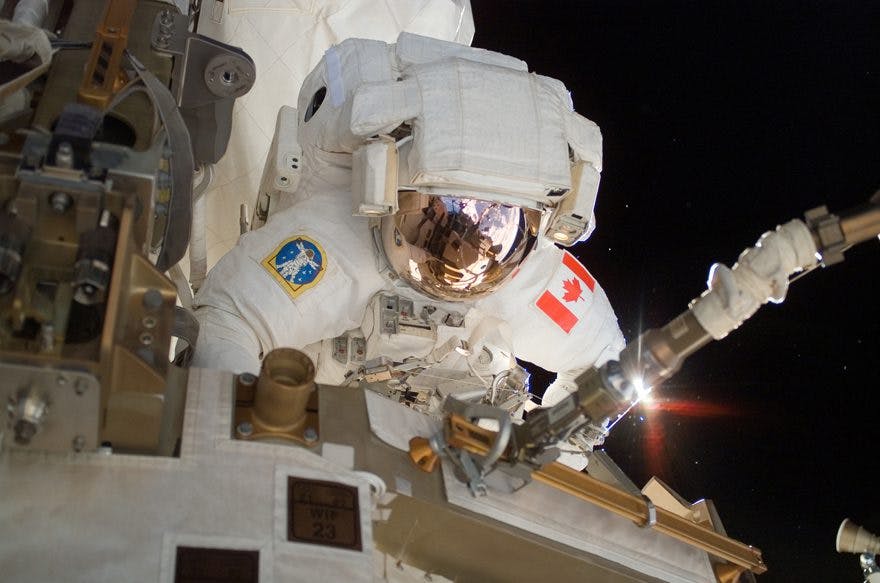 An astronaut in a white spacesuit, with a Canadian flag on the sleeve, works outside a space station in the darkness of space.