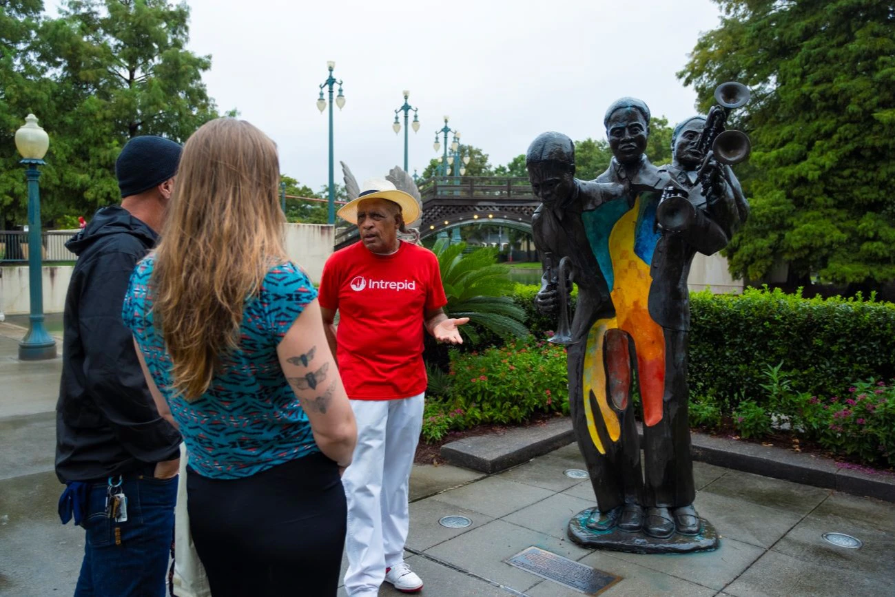 A man in a red Intrepid shirt speaks to a woman near a colorful jazz musician statue in a park with trees and lampposts.