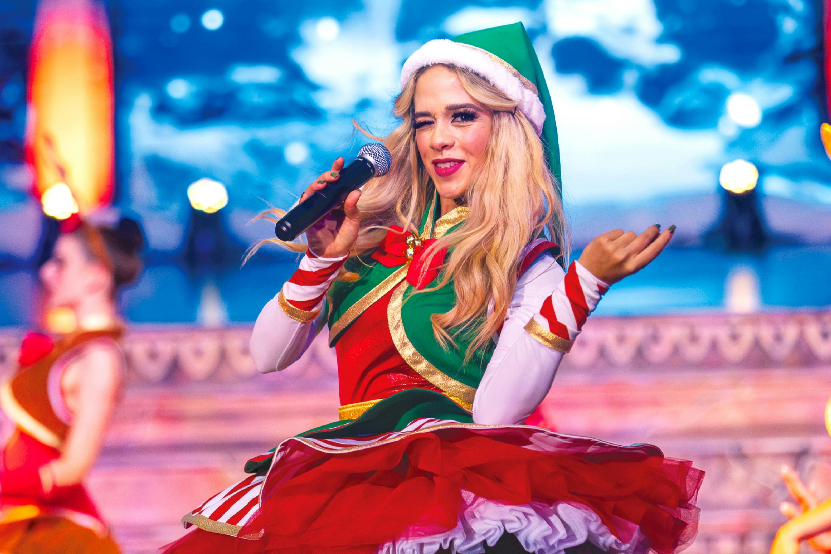 Γυναίκα με γιορτινή στολή ξωτικού τραγουδά σε μικρόφωνο επί σκηνής με χιονισμένο φόντο.