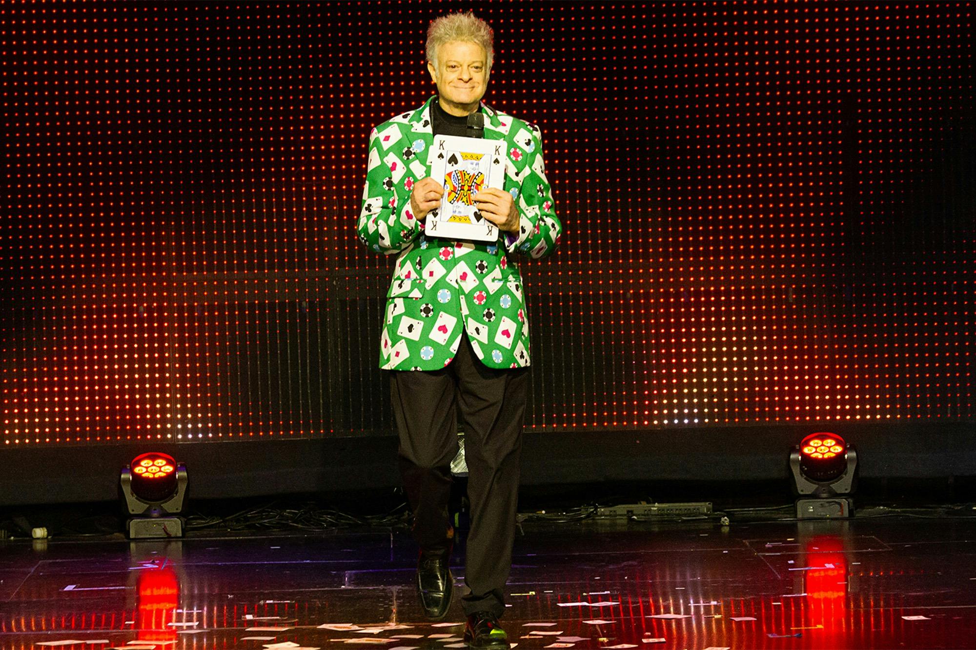 A man in a green card-themed jacket holds up a card on a stage with a red light-dotted backdrop.